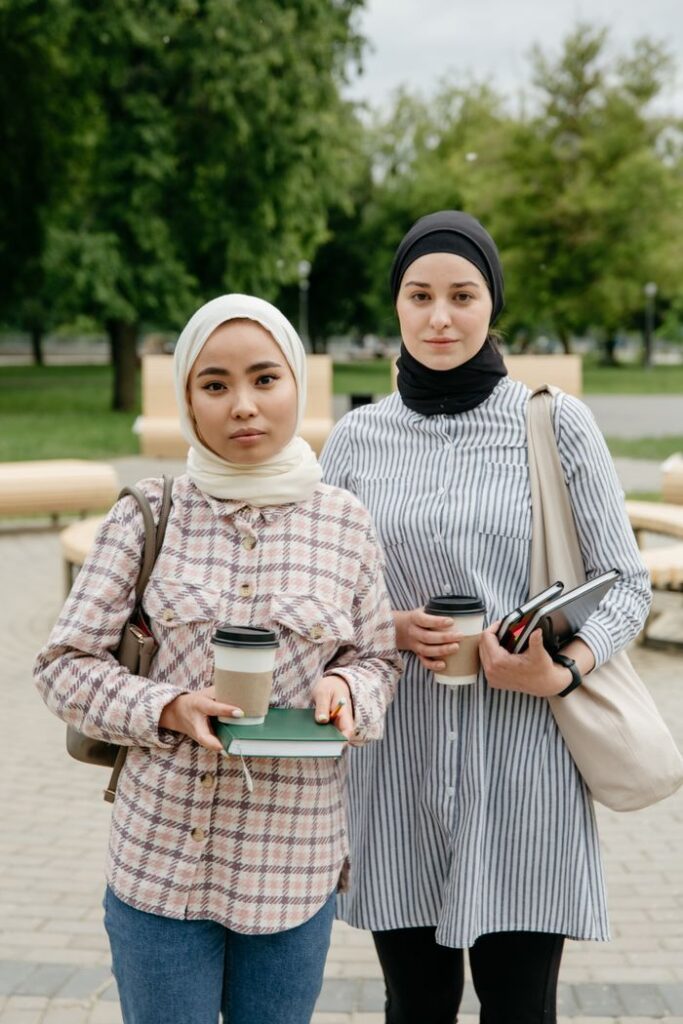 Two women in hijabs holding coffee cups and notebooks, standing outdoors.